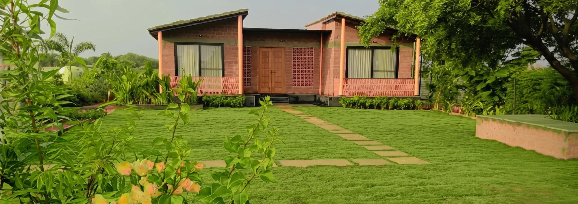 On-site photograph of the model farmhouse at The Sensory: a single-storey brick cottage with a pitched terracotta-tile roof, timber-look support columns, a covered veranda with a lattice-pattern balustrade, a carved wooden double door, and a freshly laid lawn with a stone pathway leading to the porch, palms and flowering shrubs in the foreground.